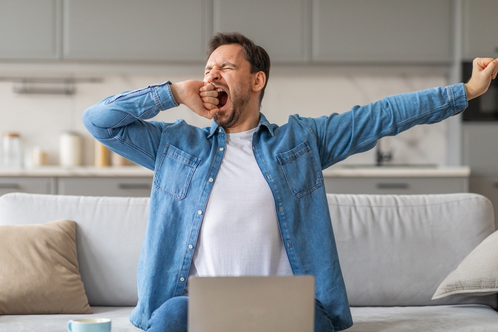 Man yawning sitting on couch in front of laptop