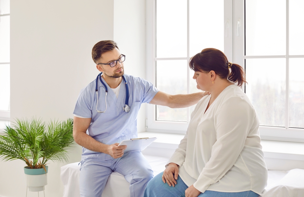 A male doctor in blue scrubs holding a clipboard and talking to a female patient in a bright clinic