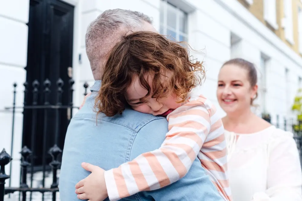 A father comforting his young child with a hug while a mother smiles in the background.