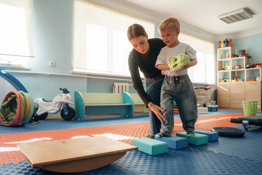 Physical therapist assisting a young boy with balance exercises on blue foam blocks.