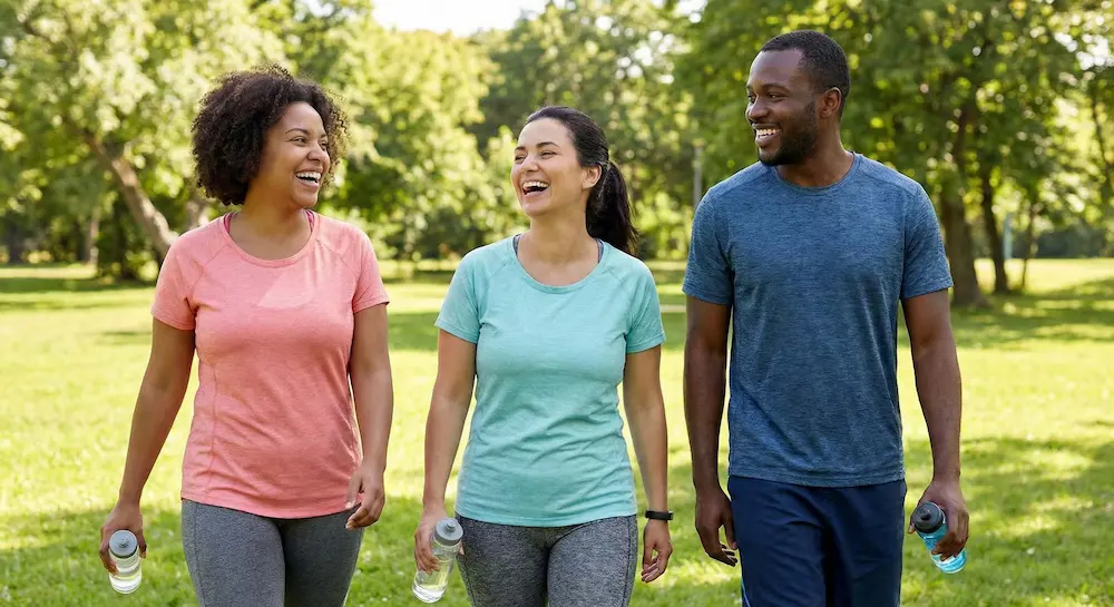 Three adults laughing while walking together in a park holding water bottles.