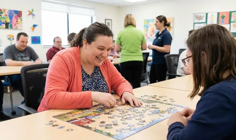 Woman smiling while working on a jigsaw puzzle with a companion in a community activity room.
