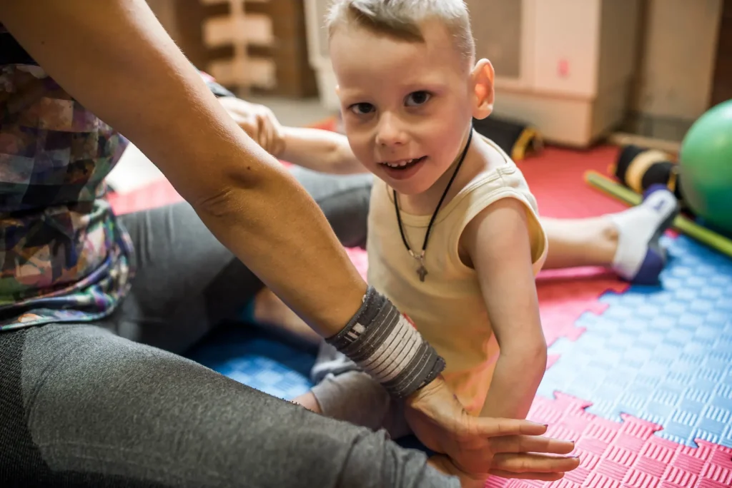 A caregiver gently holding hands with a young boy during an at-home therapy session on a colorful foam mat, supporting the behavioral and physical needs of LGS.