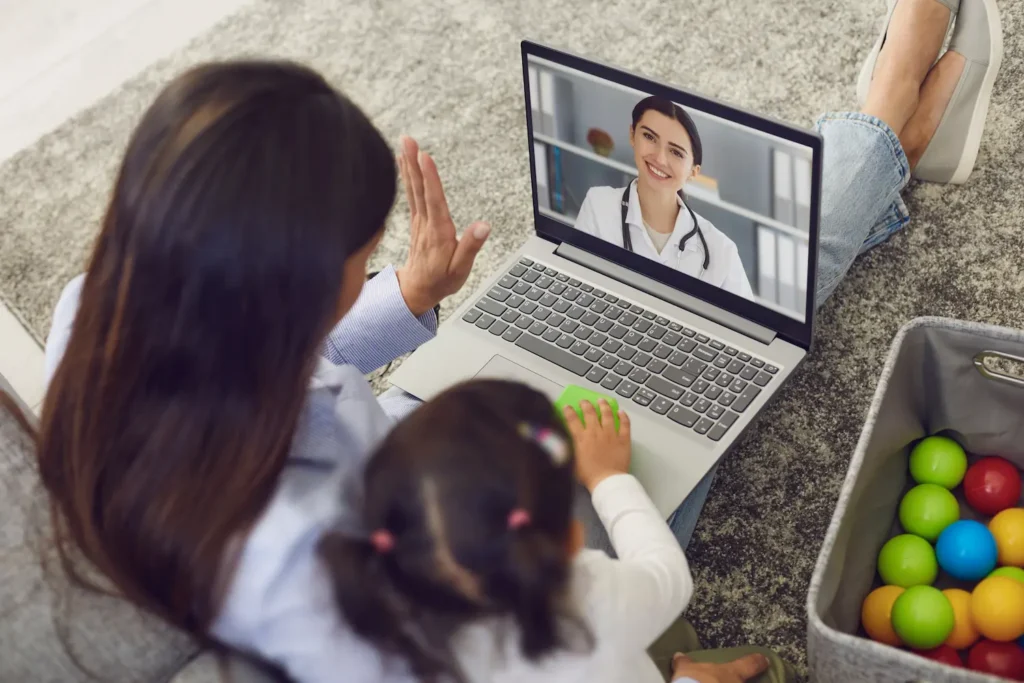 A mother and child waving to a doctor on a laptop, illustrating a virtual telehealth visit for at-home clinical trials.